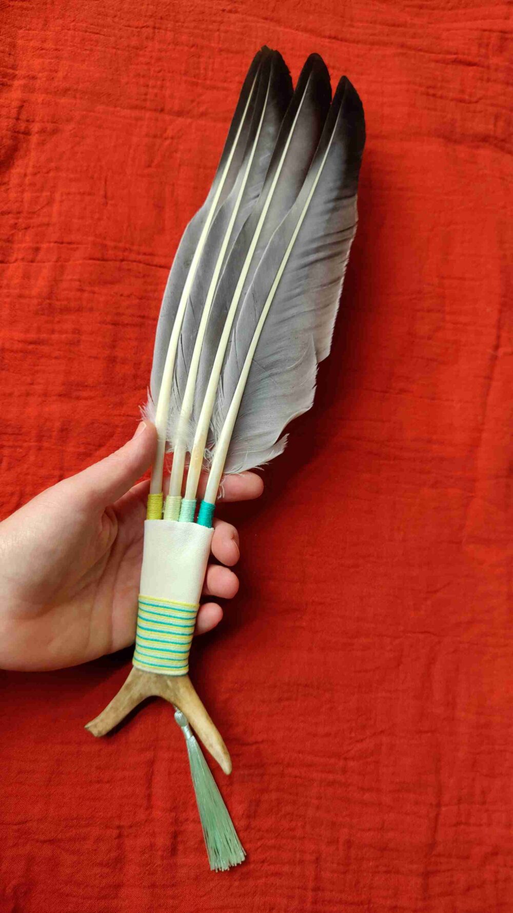 Räucherfächer aus grauen Gänsefedern und Rentierhorn, der von einer Hand gehalten wird, vor einem roten Tuch liegt.