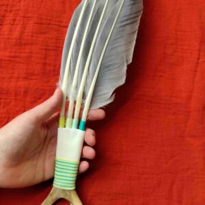 Räucherfächer aus grauen Gänsefedern und Rentierhorn, der von einer Hand gehalten wird, vor einem roten Tuch liegt.
